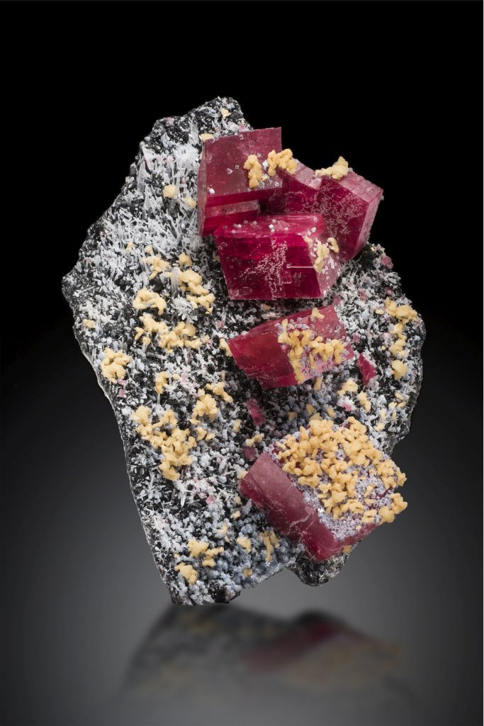 A photo of the Alma Rose rhodochrosite specimen, it is a black rock with gray and yellow crystal formations and 6 large rhodochrosite cubes. Featured on a black background.