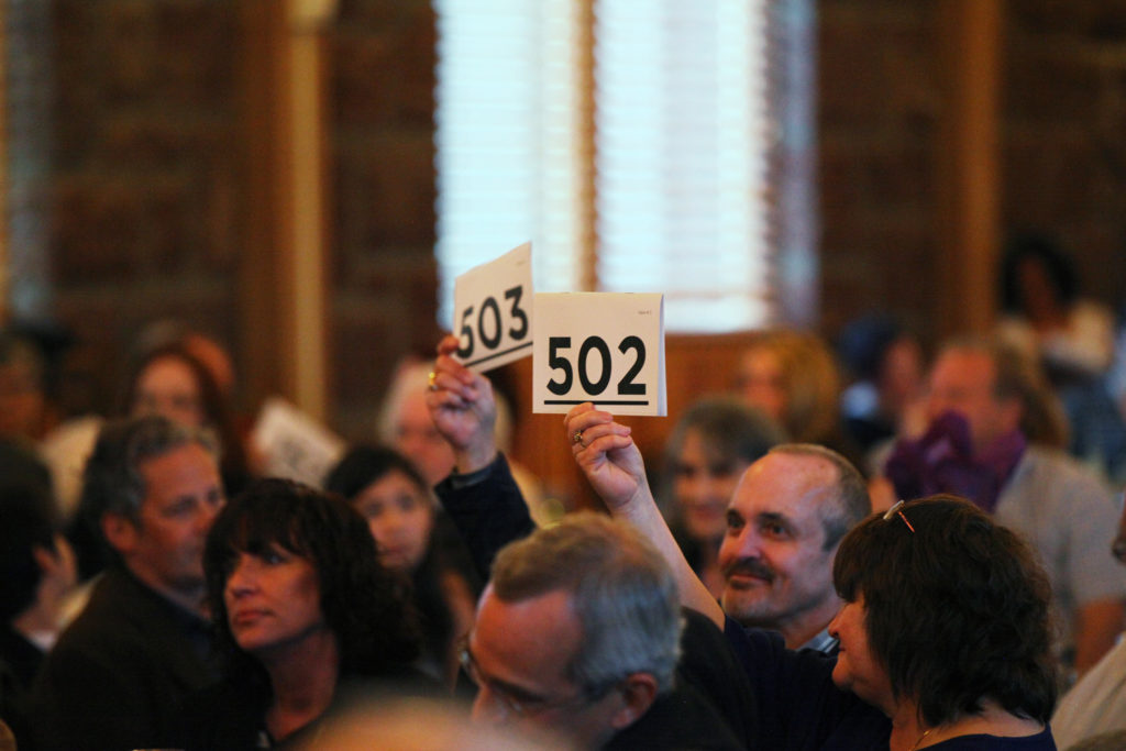 A crowd of people seated in front of a window, mostly blurred. A man and a woman are in focus, holding up auction paddles with the numbers