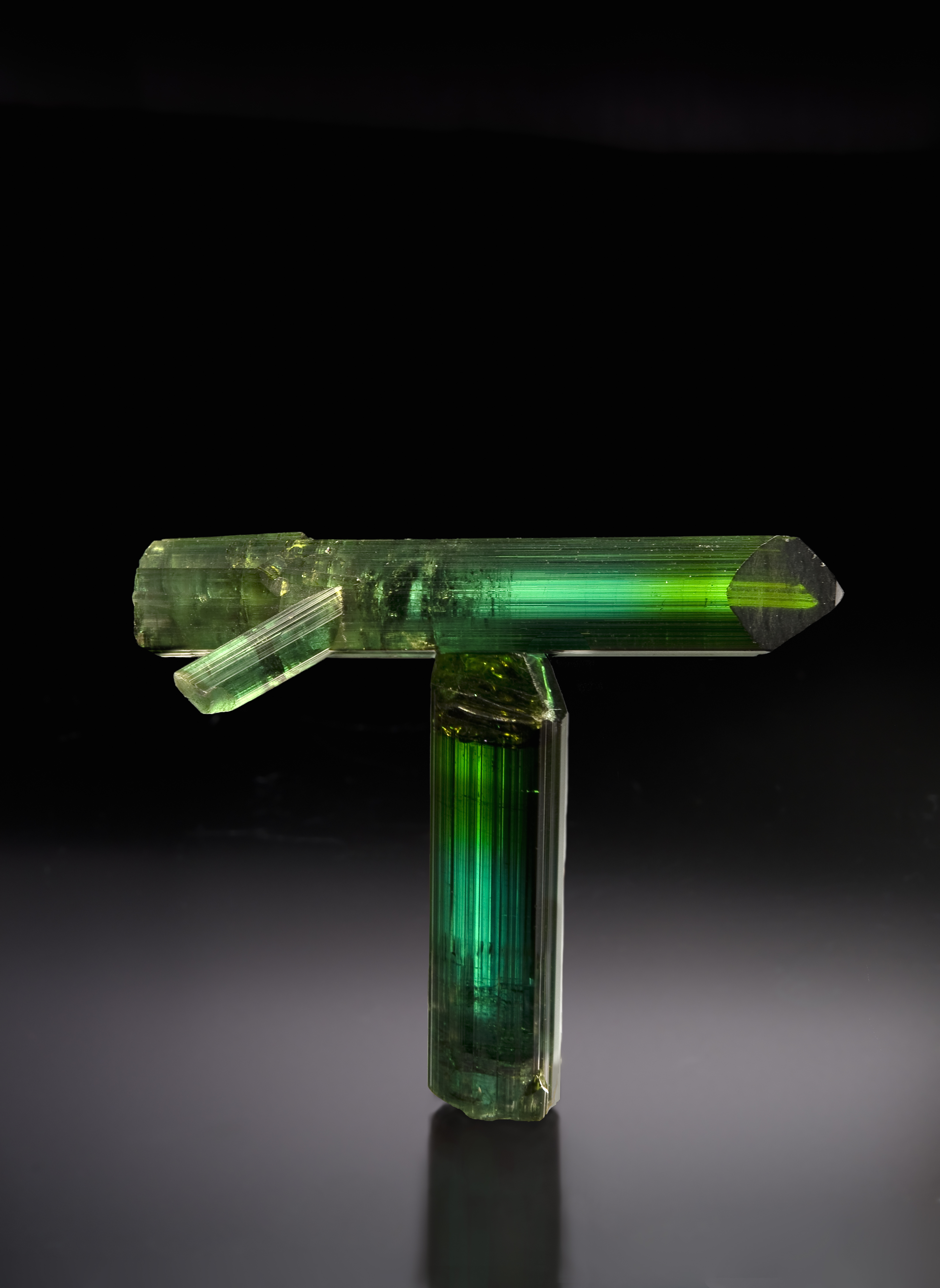 Two kelly green rounded tourmaline crystals form a capital T on a gray background. They are lit from above and reflect on the gray surface. The top crystal has a small adjoining crystal that makes it appear as if it is a rocket, about to take off to the right of the frame. 