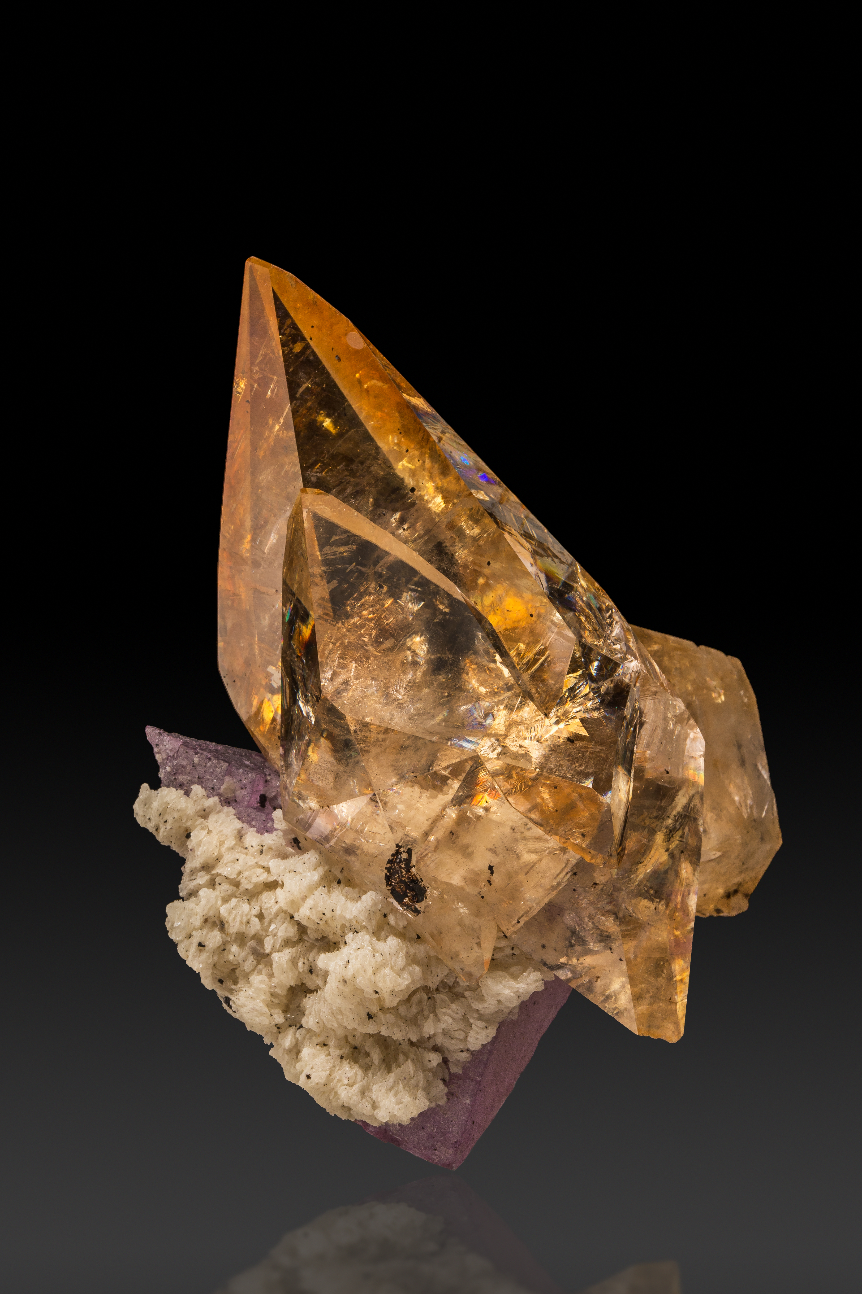 A large, clear orange calcite points to the upper left of the image. It appears to be bursting forth from a gray and purple base, barely visible on the bottom left. On a black background. 