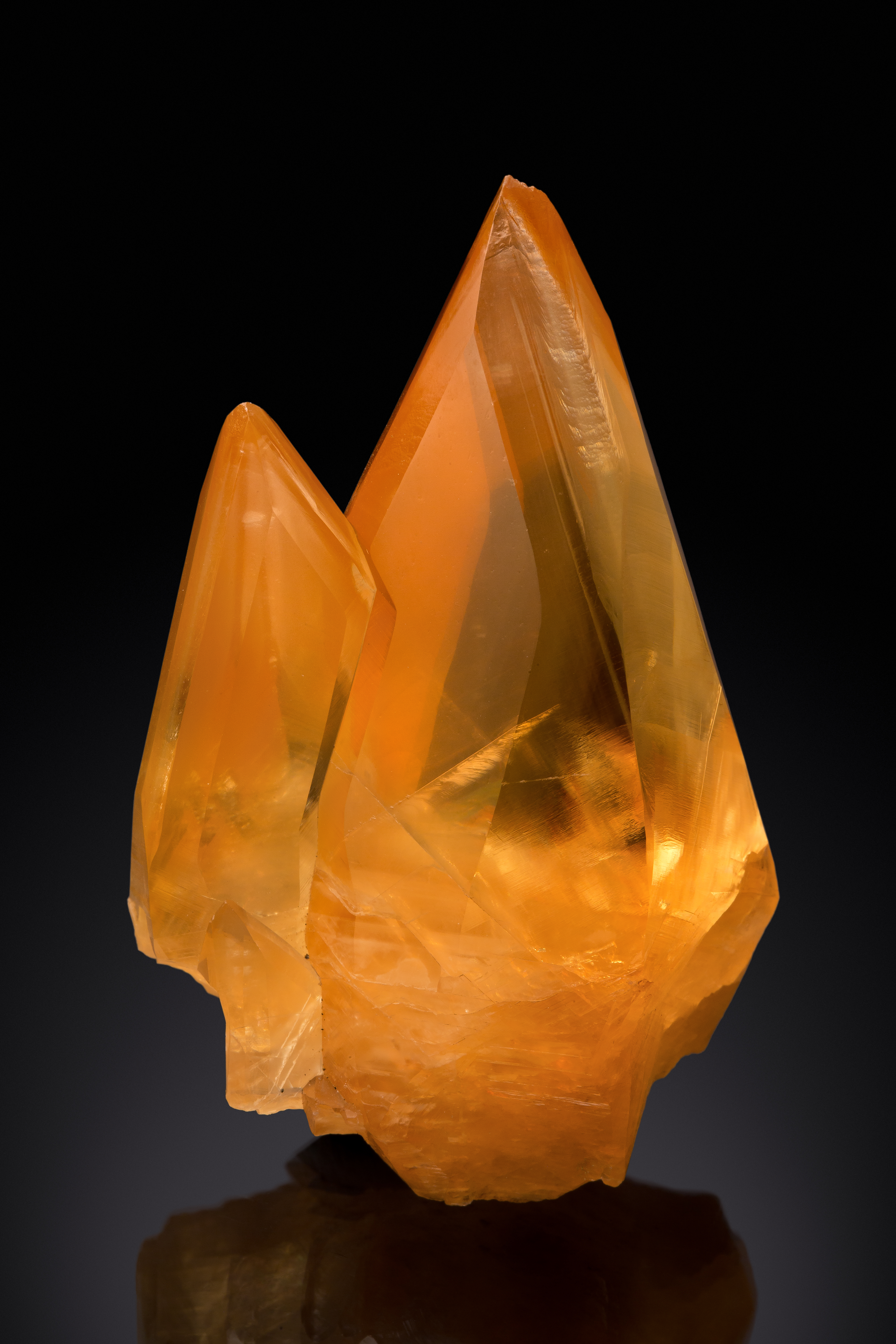 A cheerful, bright orange calcite crystal with two points extending upward to the top of the frame. Wider at the bottom with two points at the top, it is reminiscent of a flame against a black background. 