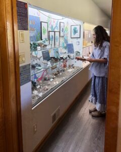 Intern Paulina in front of a long display case with a variety of minerals and signage on the top two sleeves. A sand tray spans the bottom shelf with shells and more minerals. Artwork of mermaids and sea life hang on the back wall.